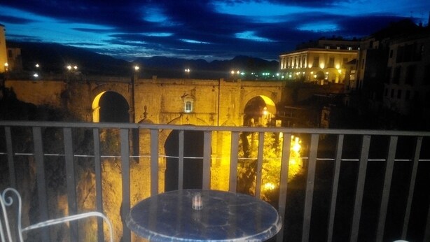 Een balkon met een ronde tafel en twee stoelen, met uitzicht op een historische stenen brug met bogen en verlichte gebouwen op de achtergrond.