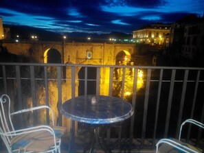 Un balcón con una mesa redonda y dos sillas, con vistas a un puente de piedra histórico con arcos y edificios iluminados al fondo.