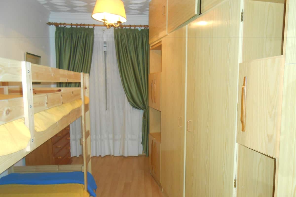 Een slaapkamer met stapelbed, een houten vloer, een bed met een blauwe en gele deken, een houten kledingkast en een hanglamp.