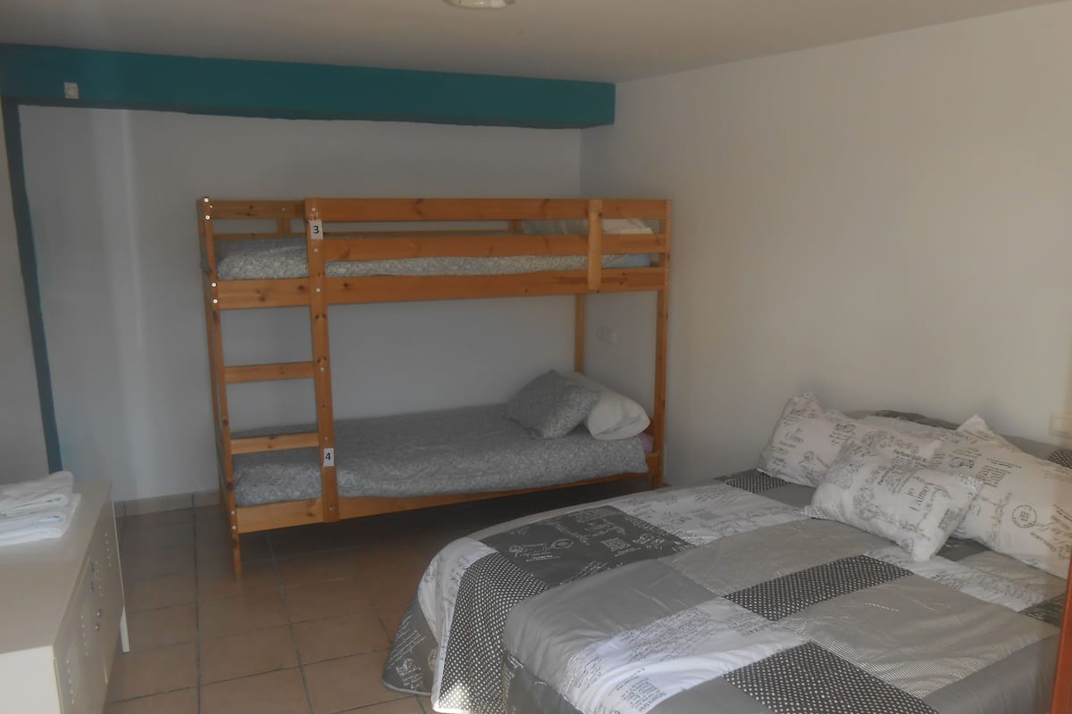 Een stapelbed met een onderbed en een eenpersoonsbed, beide met beddengoed met een patroon. Er ligt een betegelde vloer en een witte muur met blauwe rand.