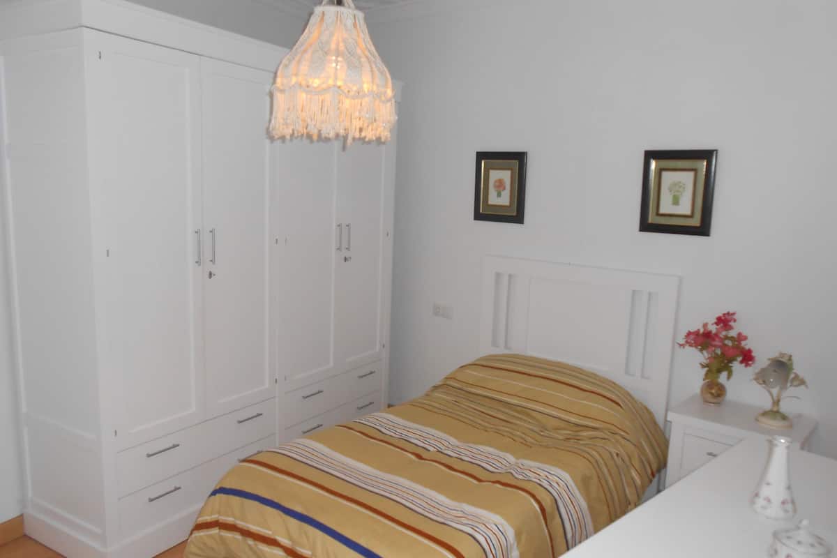 Een slaapkamer met een bed, een kledingkast, een nachtkastje, een vaas met bloemen en een kroonluchter.