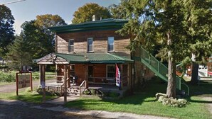 A rustic log cabin with a porch, a small attached shed, and a flag. - The School House Inn (Altmar)