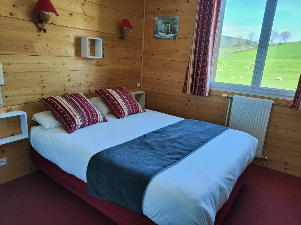Ein Zimmer mit Holzwänden, einem Bett, zwei Wandlampen, einem Gemälde und einem Fenster mit Blick auf ein grünes Feld.