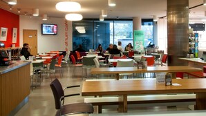 A modern cafe with wooden tables and red chairs, a counter with a cash register, and a large pillar. - Petros Court (Canterbury)