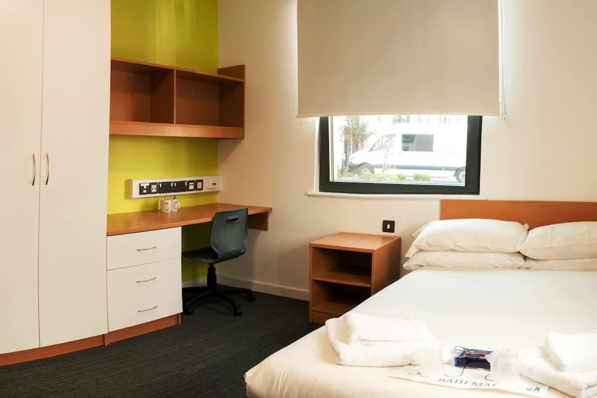A hotel room with a bed, a desk, a chair, a wardrobe, and a window with a view of the outside.