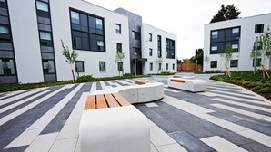 A modern outdoor area with a geometric tile pattern, white and gray seating, and a building with large windows and a flat roof. - Petros Court (Canterbury)