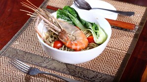 A bowl of noodles with shrimp, vegetables, and bok choy on a woven placemat.