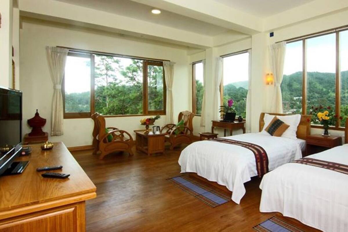 Habitación de hotel con dos camas, televisión, muebles de madera y amplias ventanas con vistas a la montaña.