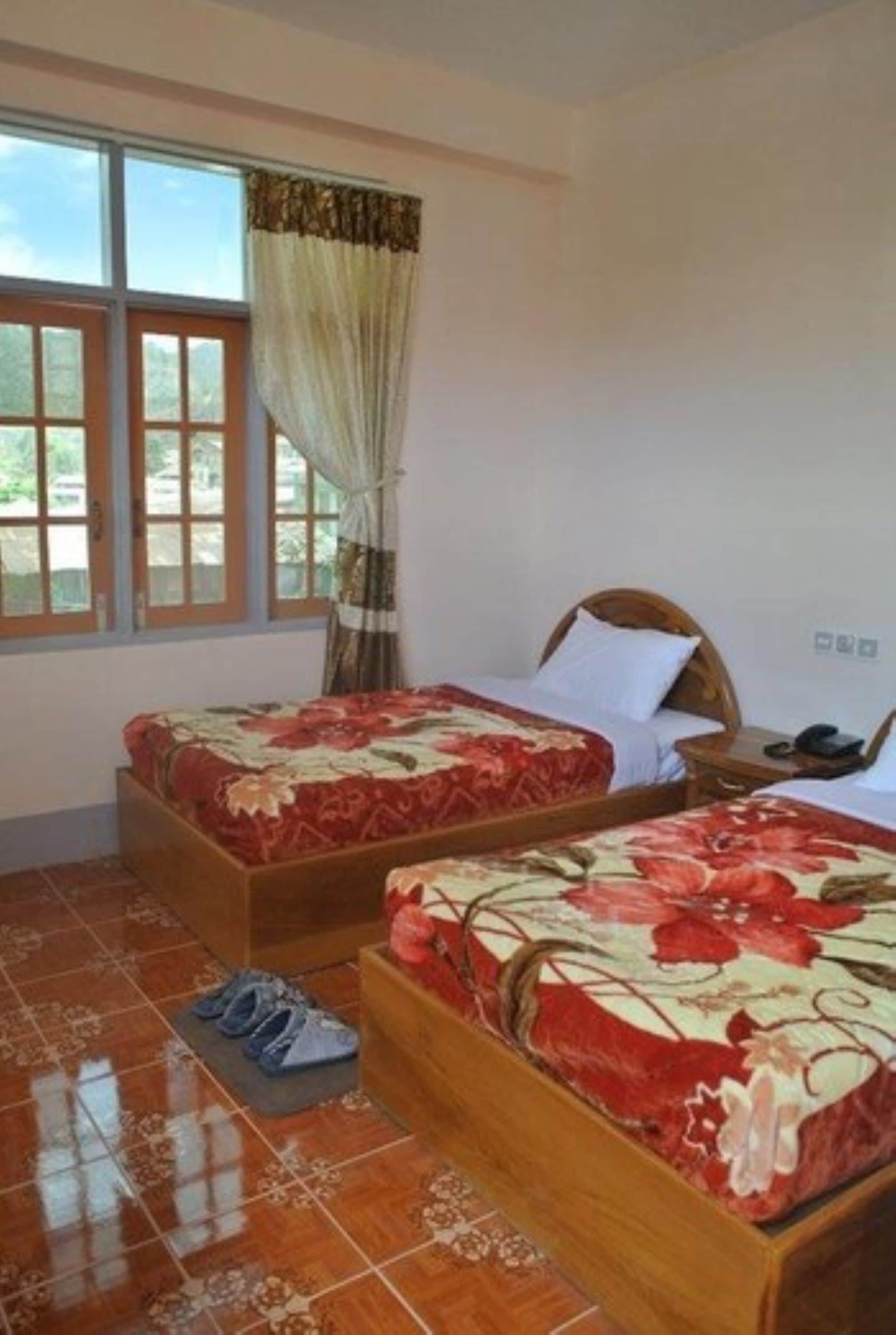 A room with two wooden beds with floral bedding, a window with curtains, and a pair of shoes on the floor.