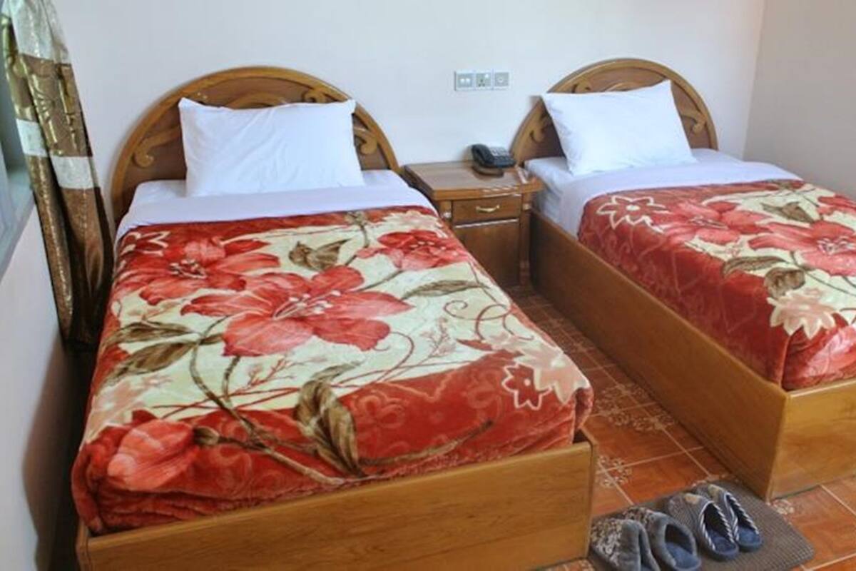 Two wooden beds with floral bedding, a bedside table with a phone, and a pair of slippers on the floor.