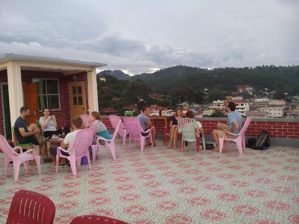 Un grupo de personas sentadas en sillas de plástico alrededor de una mesa en una terraza con suelo estampado, con vistas a un paisaje montañoso.