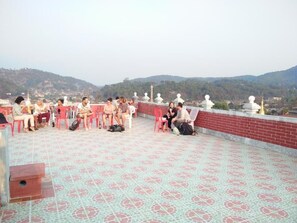 A rooftop terrace with people seated on plastic chairs, a patterned floor, and a view of hills in the background.