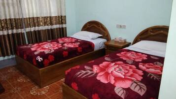 A room with two wooden beds, each with a floral bedspread. There is a nightstand with a lamp and a box on it. The room has a tiled floor and a curtain on the window.