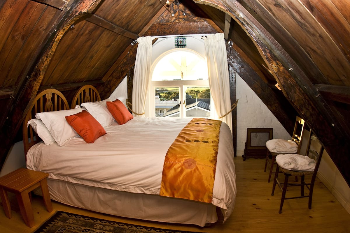 A cozy attic bedroom with a large bed, wooden chairs, and a view of the outside.