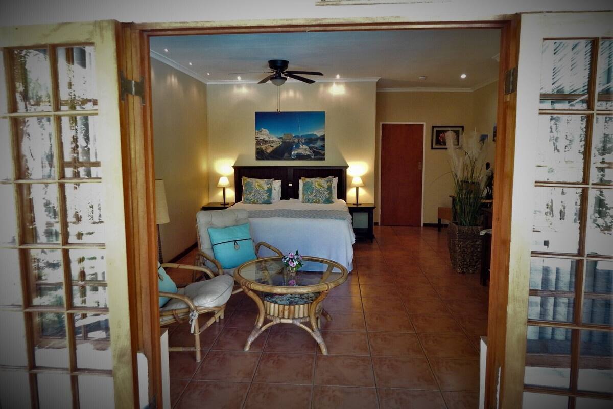 A hotel room with a bed, a ceiling fan, a nightstand, a table with flowers, and a view of the outside.