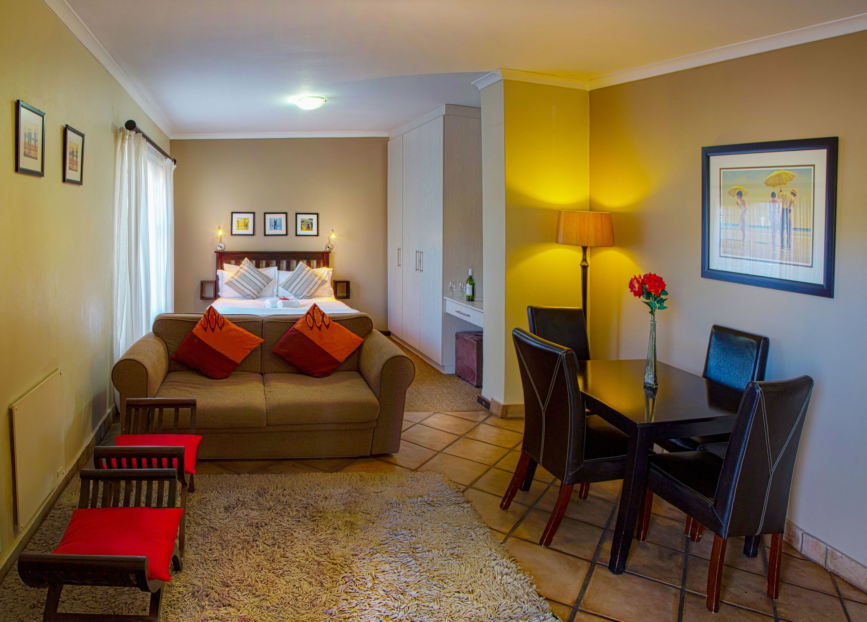 A hotel room with a bed, a sofa, a dining table with chairs, and a small table with a vase of flowers.