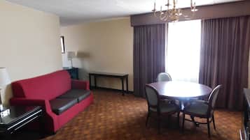 A hotel room with a red sofa, a dining table with chairs, a chandelier, a lamp, and a window with curtains.