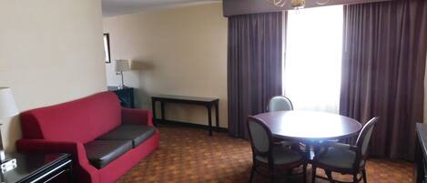 A hotel room with a red sofa, a dining table with chairs, a chandelier, a lamp, and a window with curtains.