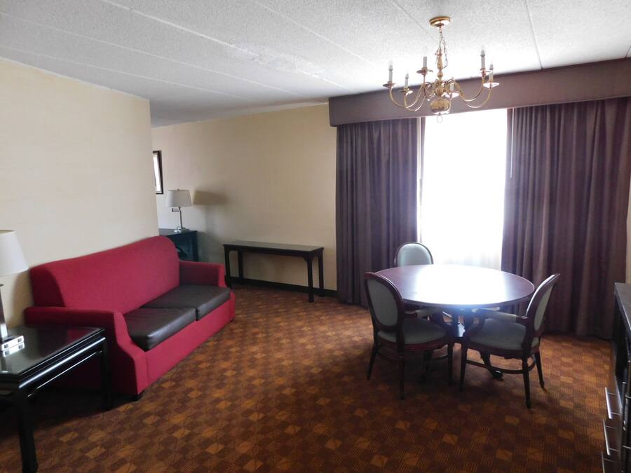A hotel room with a red sofa, a dining table with chairs, a chandelier, a lamp, and a window with curtains.