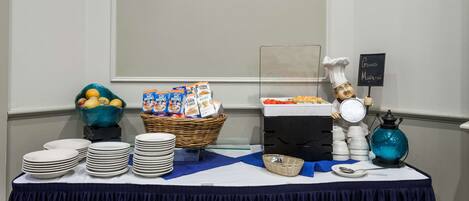 Una mesa de buffet con platos apilados, un bol de fruta, cajas de cereales y un cartel que indica una estación de café.