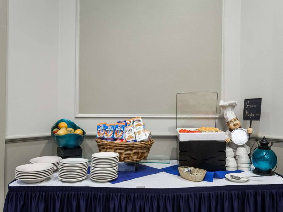 Una mesa de buffet con platos apilados, un bol de fruta, cajas de cereales y un cartel que indica una estación de café.