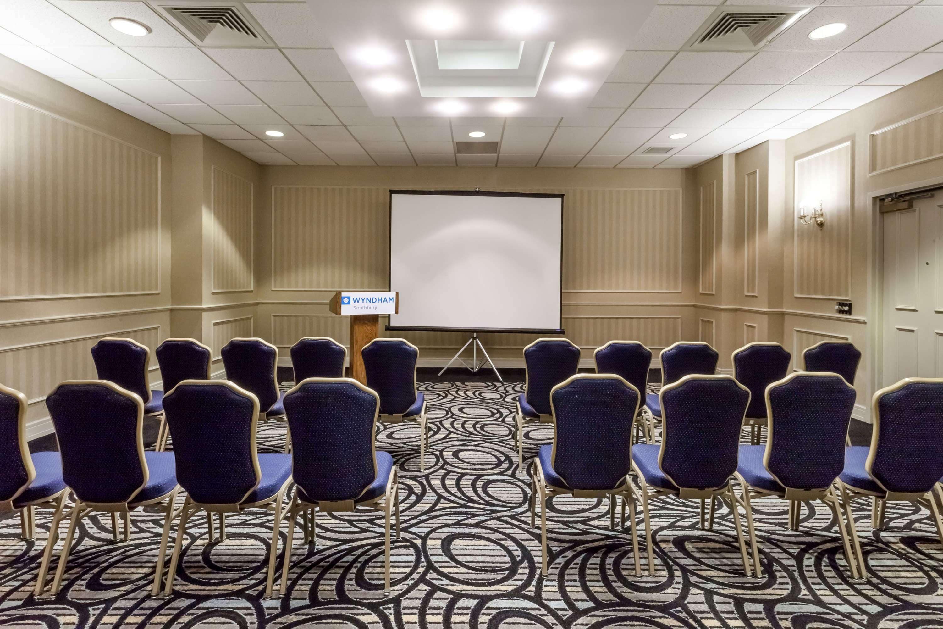 A conference room with rows of blue chairs, a large projection screen, and a carpeted floor.