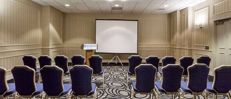A conference room with rows of blue chairs, a large projection screen, and a carpeted floor.