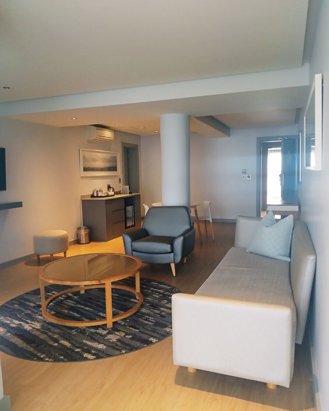 Ein modernes Wohnzimmer mit Sofa, Sessel und rundem Couchtisch. Es gibt einen Essbereich mit Tisch und Stühlen sowie eine Küchenzeile mit Arbeitsfläche und Schränken.