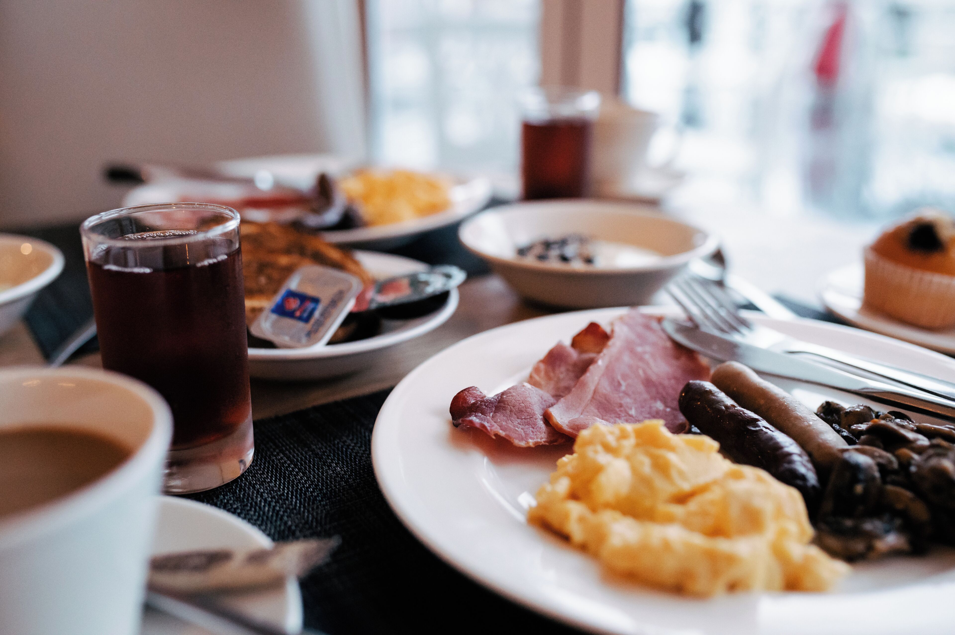 A breakfast setting with plates of eggs, bacon, sausages, and beans, a glass of dark beverage, and a muffin.