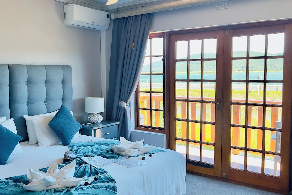 A hotel room with a large bed, a bedside table with a lamp, and a view of a wooden deck and greenery through a glass door.