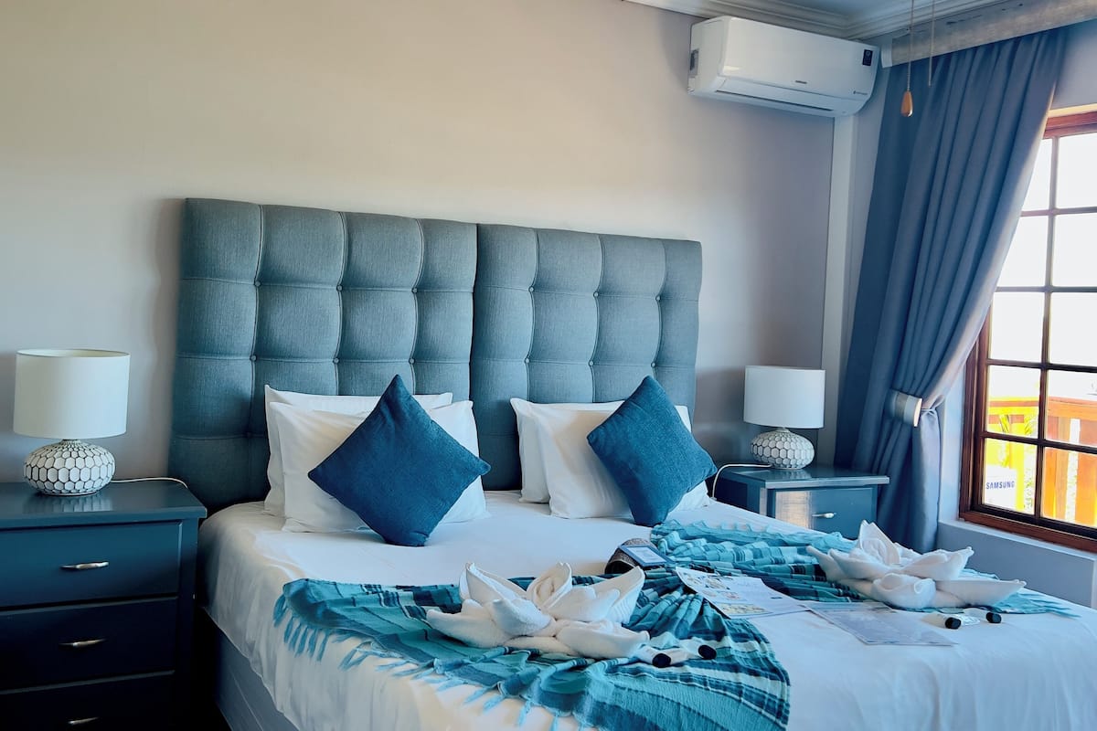 A hotel room with a tufted headboard, a bed with white and blue bedding, two bedside tables with lamps, a chair, and a window with curtains.