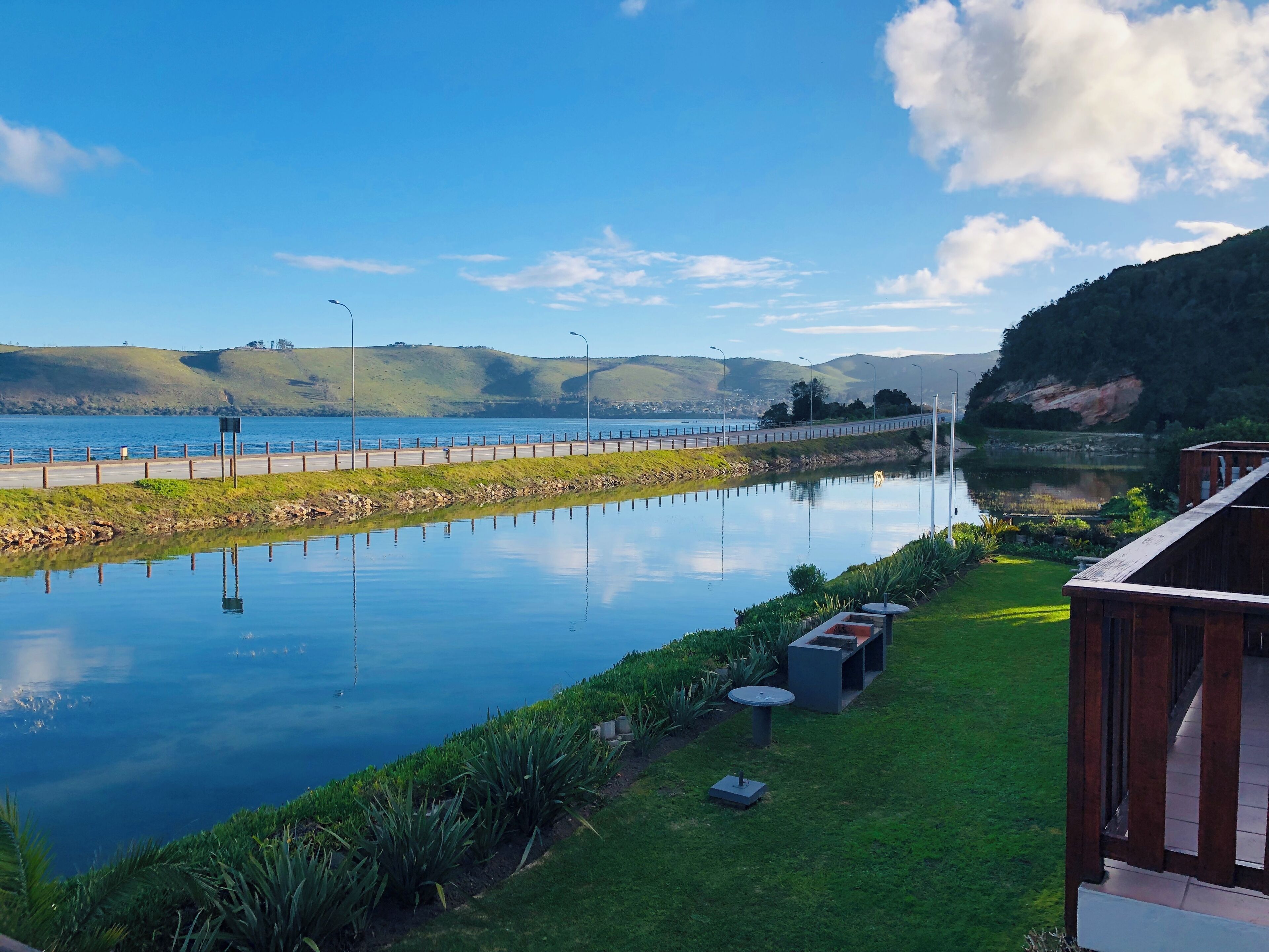 A serene lakeside view with a wooden deck and lounge chairs, a paved walkway, and a distant road.