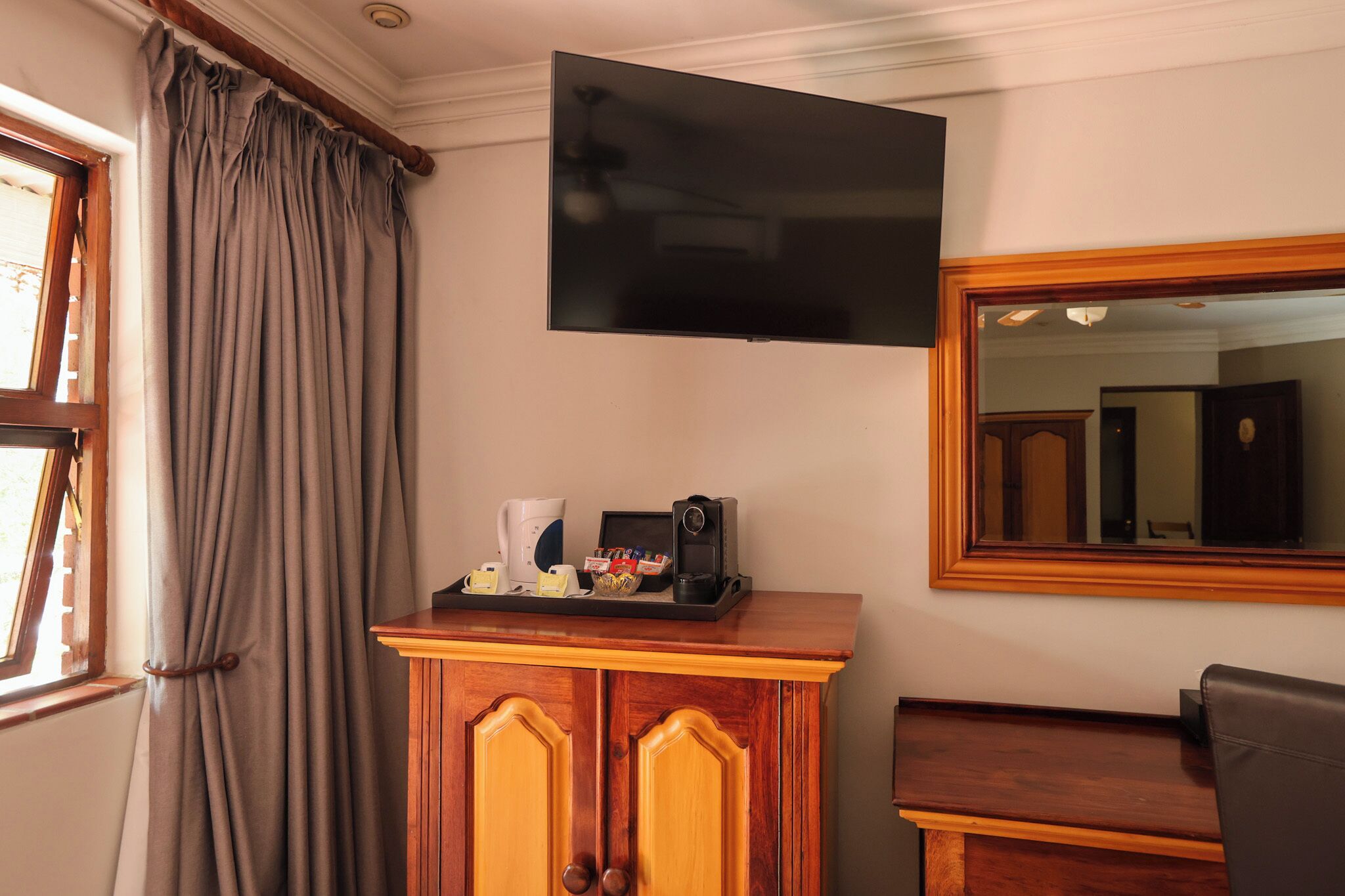 A hotel room with a flat-screen TV mounted on the wall, a wooden cabinet with a tray of coffee-making items, and a large mirror.