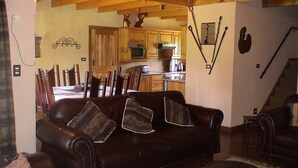 A cozy living room with a brown leather sofa, wooden dining chairs, a kitchen in the background, and a staircase to the right. - Champagne Haven (Cathkin Park)