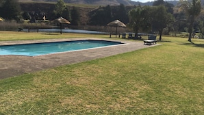 A rectangular swimming pool with a paved deck, surrounded by a grassy area with a few trees and a thatched-roof structure. - Champagne Haven (Cathkin Park)