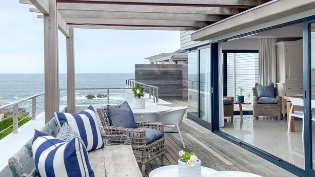 A modern outdoor deck with wicker furniture, a white table, and a view of the ocean. - Le Paradis Penthouse Apartment (Hermanus)