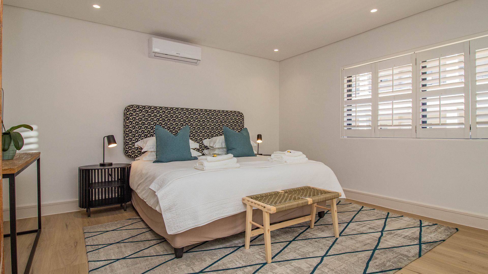 Une chambre moderne avec une tête de lit à motifs, une table de chevet avec une lampe, un banc et une fenêtre avec des stores.