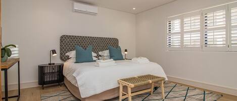 Une chambre moderne avec une tête de lit à motifs, une table de chevet avec une lampe, un banc et une fenêtre avec des stores.