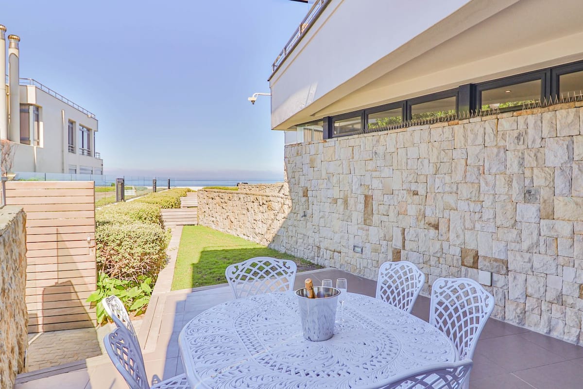 A modern outdoor patio with a stone wall, a round table with chairs, and a view of the sea.