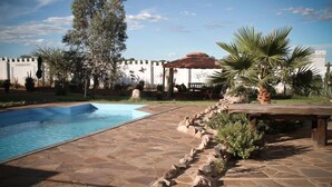 Une piscine aux bords recouverts de pierres, entourée d’un jardin agrémenté de palmiers et d’une pergola.