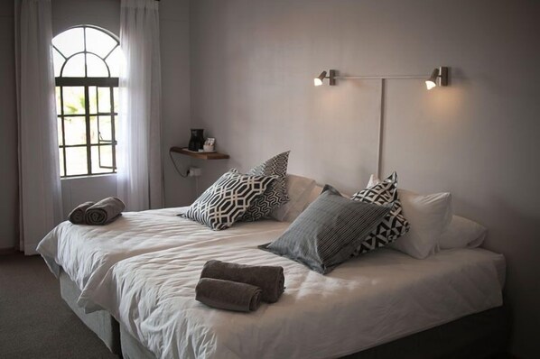 A double bed with patterned pillows, a bedside table with a lamp, and a window with curtains. - Sweet Thorn Retreat Airport Lodge (Windhoek)