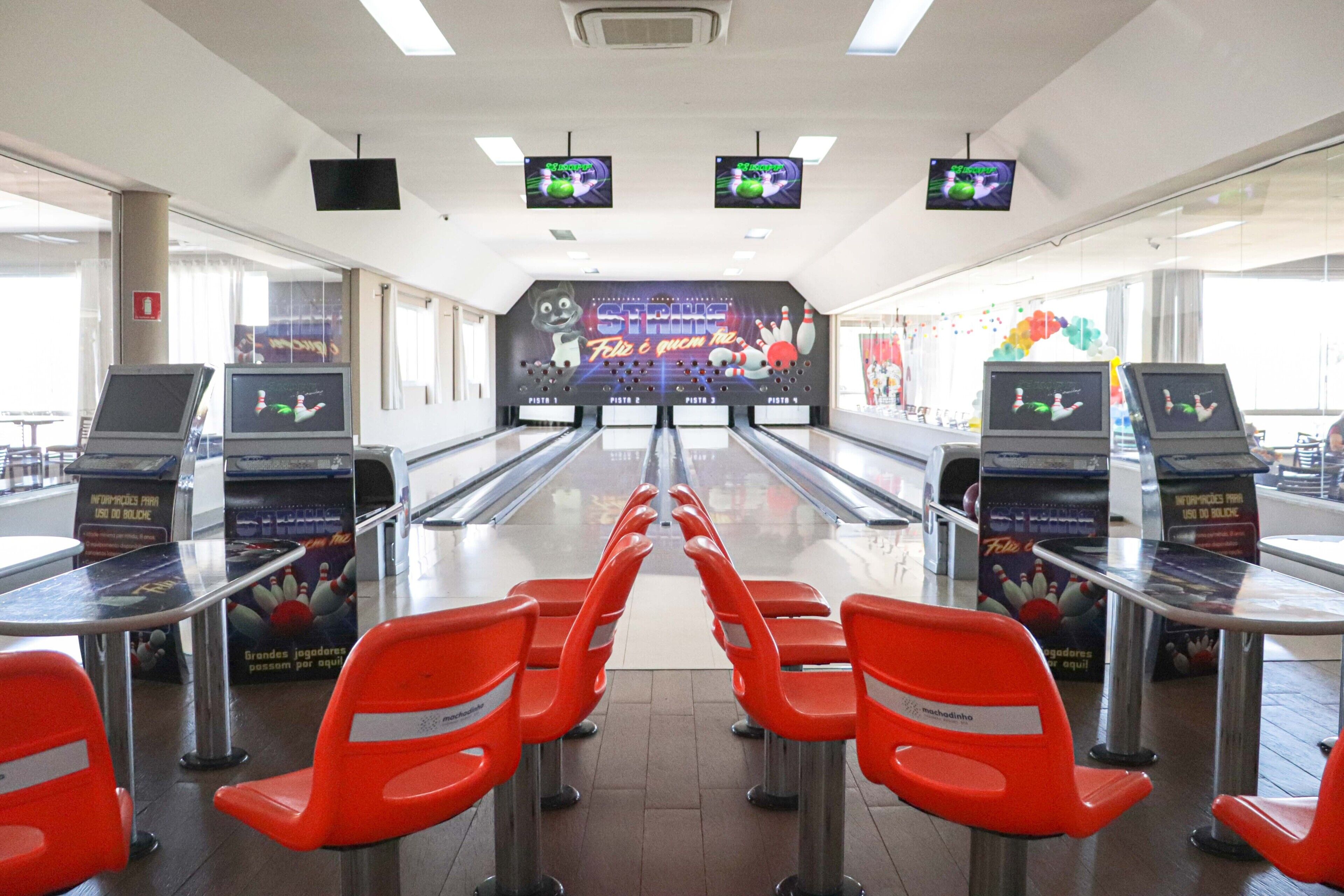 Uma pista de boliche com várias pistas, cadeiras laranjas e telas exibindo jogos de boliche.