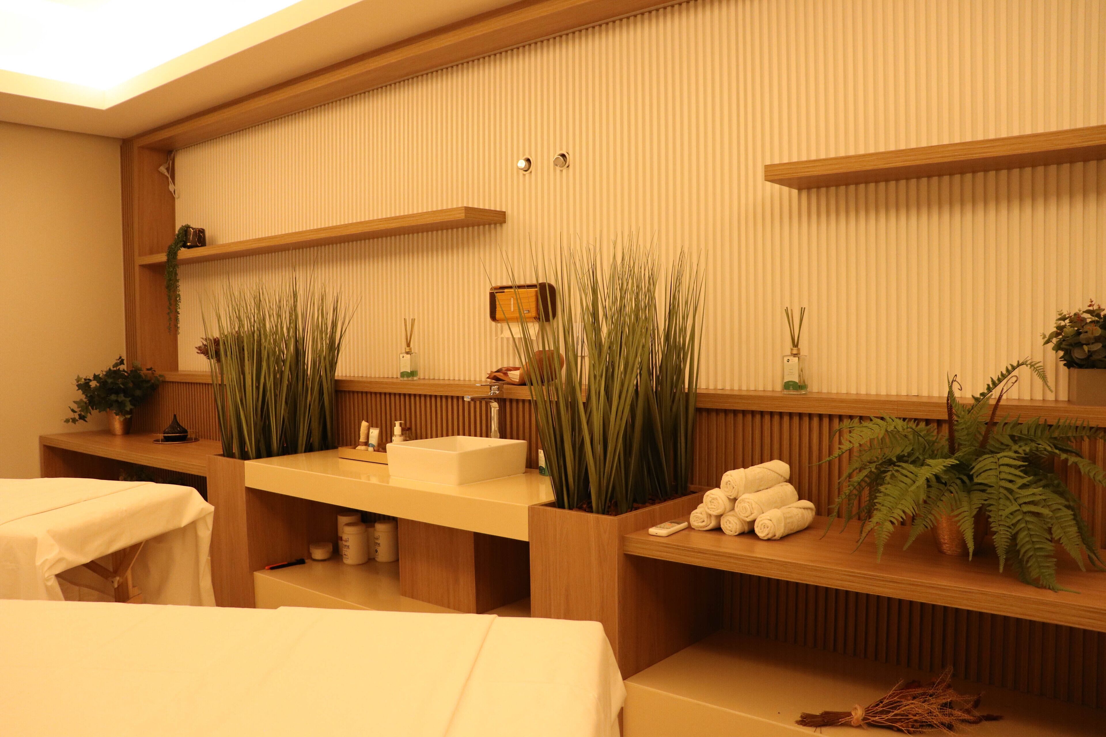 Habitación de hotel con tocador de madera, lavamanos y espejo. Hay dos plantas grandes en macetas, un estante de madera con toallas y una cama con cubrecama blanco.
