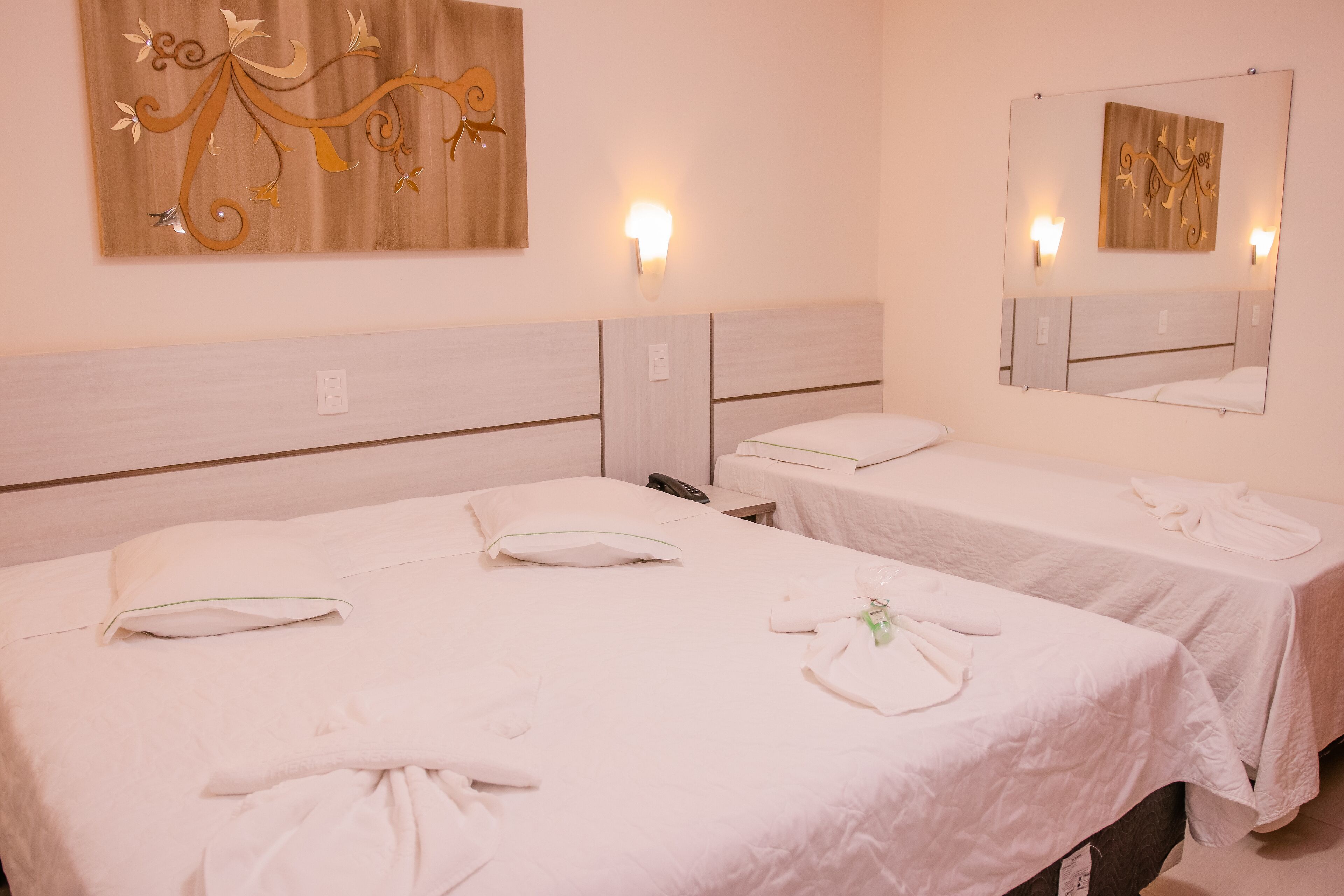 Habitación de hotel con dos camas, cada una con cubrecama blanco, almohadas y toallas. Se observan lámparas fijadas a la pared y un espejo grande sobre las camas.