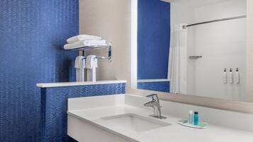 Un baño moderno con un lavamanos blanco, grifo cromado y un espejo arriba. Se observa un perchero con toallas dobladas y una zona de ducha en el reflejo.