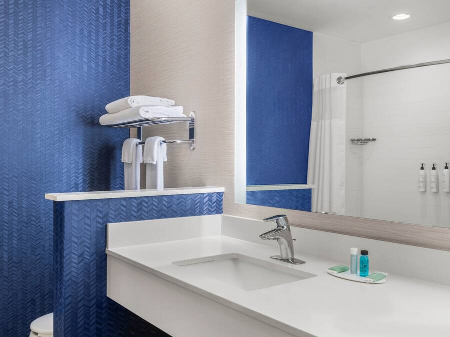 Un baño moderno con un lavamanos blanco, grifo cromado y un espejo arriba. Se observa un perchero con toallas dobladas y una zona de ducha en el reflejo.