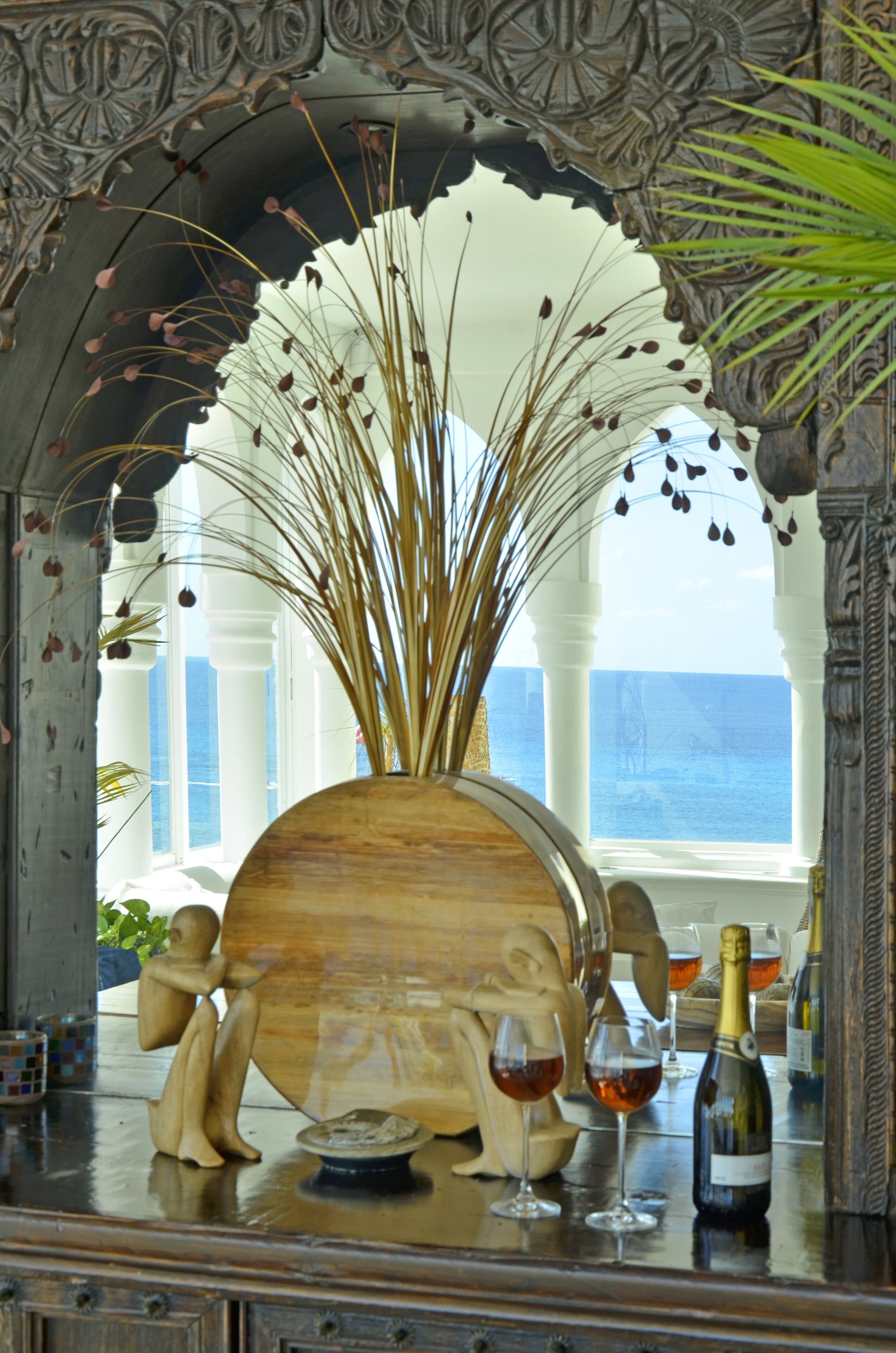 A wooden sculpture with a fan of dried plants, two wine glasses, and a bottle of wine on a reflective surface.