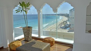 A room with a large window overlooking a beach, a wicker plant, and two ottomans.