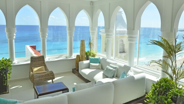 A room with a white sofa, armchairs, and a view of the ocean through arched windows.