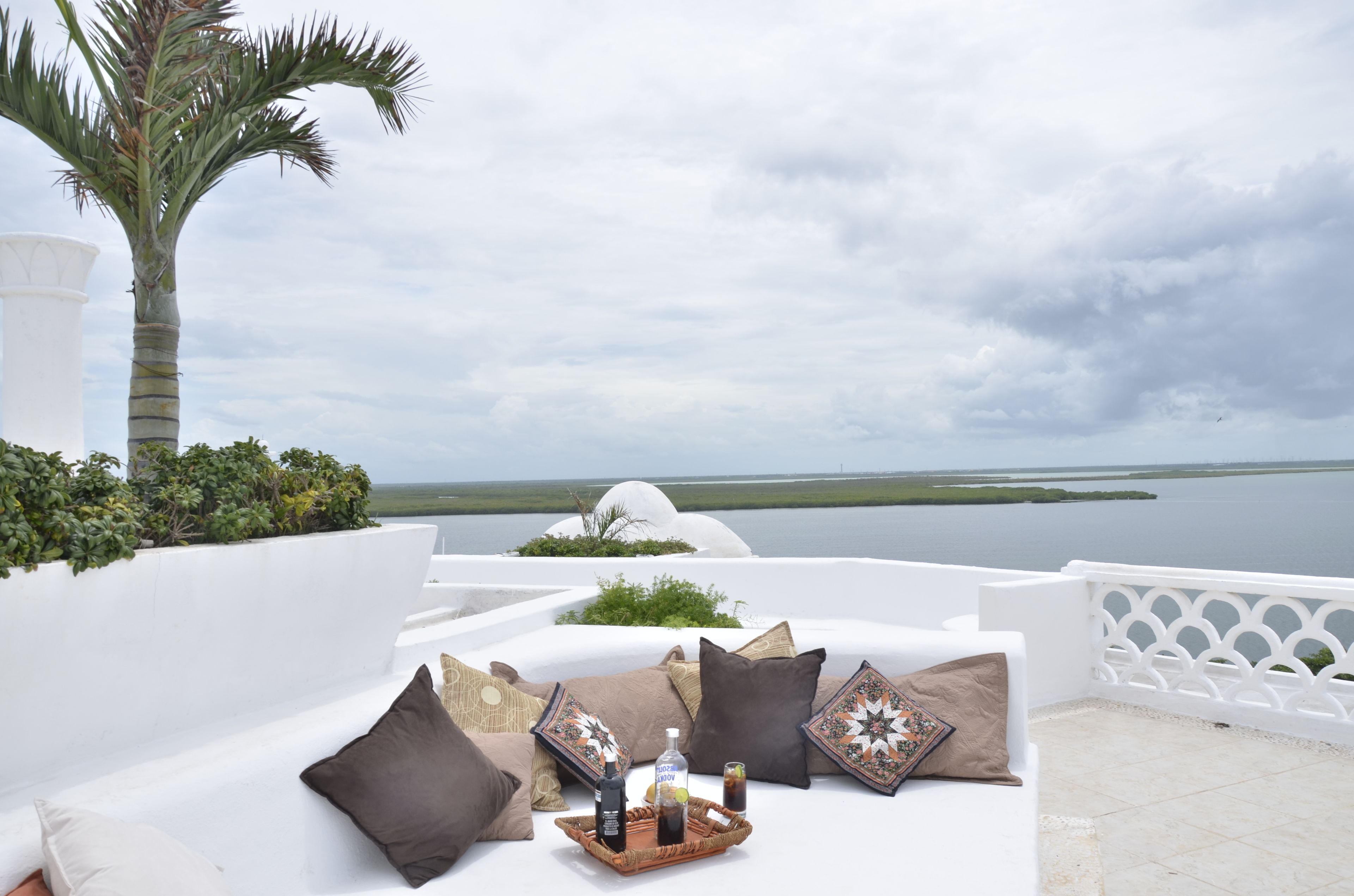 A white outdoor seating area with cushions and a tray of drinks, overlooking a body of water and palm trees.